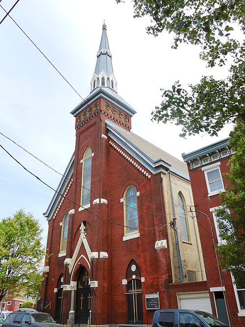 A red brick Gothic Revival church with a tall steeple and arched stained glass windows, an attached rowhouse alongside.