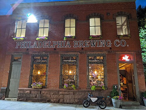 A two storey red brick building at night, PHILADELPHIA BREWING CO. painted across the upper storey above barred storefront windows.