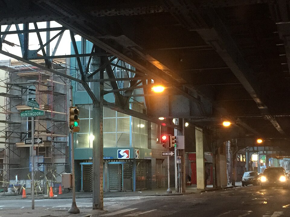 Street signs for Kensington Avenue and Huntingdon Street at a corner under the dark steel elevated superstructure, storefronts and a traffic light below.