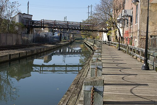 A wooden towpath along the canal, brick mill buildings on one side and an overhead pipe bridge crossing the water.