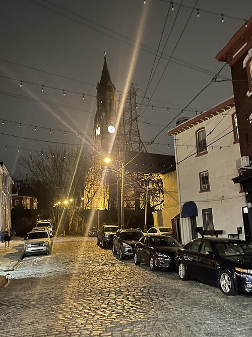 A cobblestone street lit by string lights and a streetlamp, cars parked along the curb toward an illuminated church tower.
