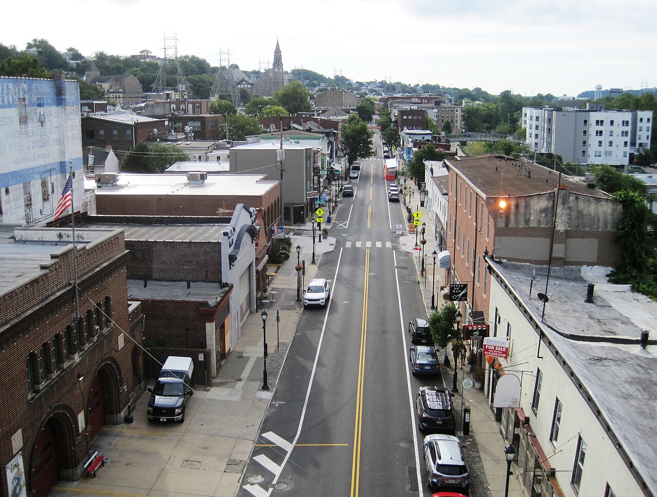 An elevated view down Main Street's storefronts toward a stone church spire, with rowhouses climbing the hillside behind on both sides.