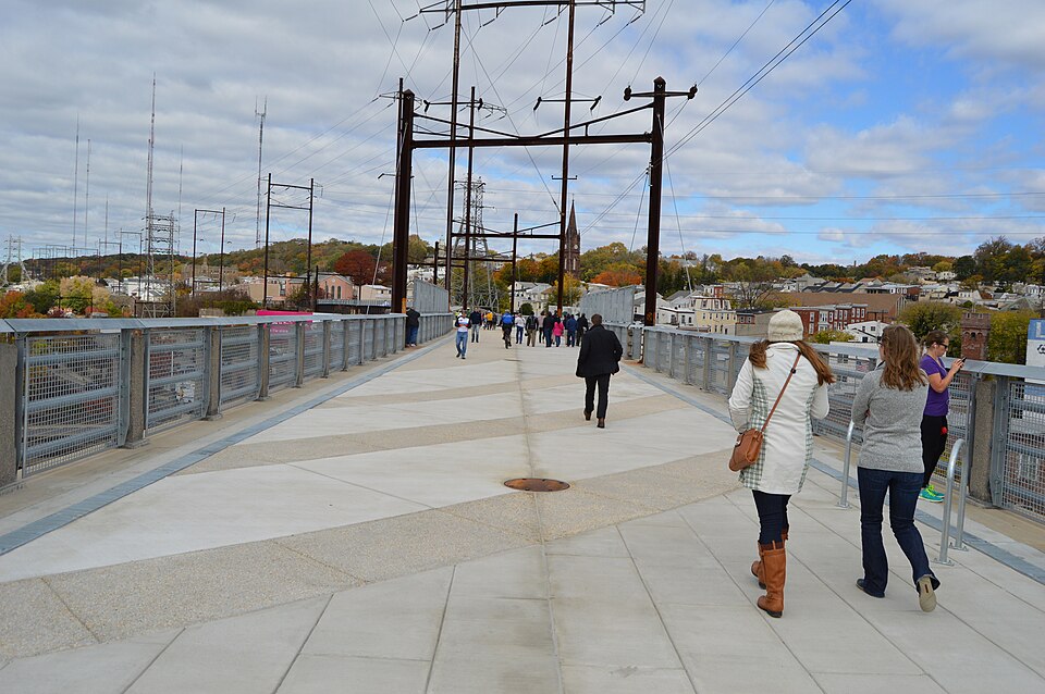 Pedestrians walking the Manayunk Bridge's paved deck between metal railings, power lines overhead.