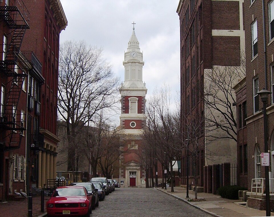 The white steeple and red brick tower of St. Augustine Church rising at the end of New Street, brick rowhouses lining both sides of the cobblestone roadway.