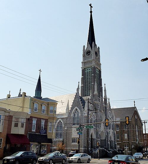 The stone Gothic Revival tower of Our Lady Help of Christians at Allegheny and Gaul, closed as a parish since 2018 but still standing beside brick storefronts in this 2021 photograph.
