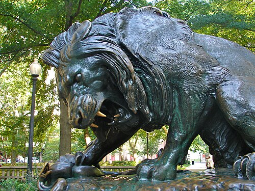A dark bronze statue of a lion crushing a serpent, on the square's own paths under the plane trees.