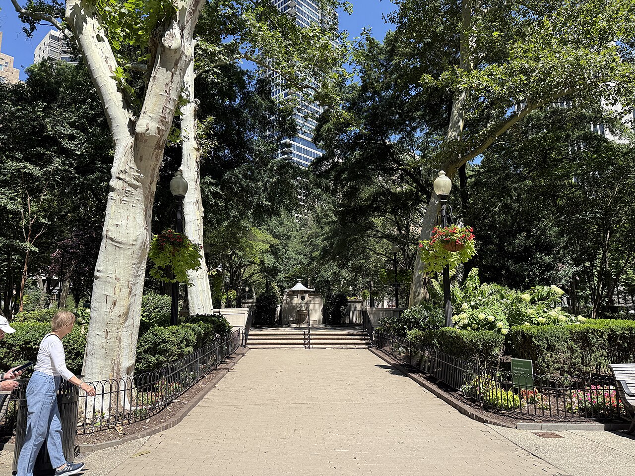 The paved diagonal walkway at the square's southeast corner, plane trees, a lantern and stone edging along the path, looking northwest into the square.