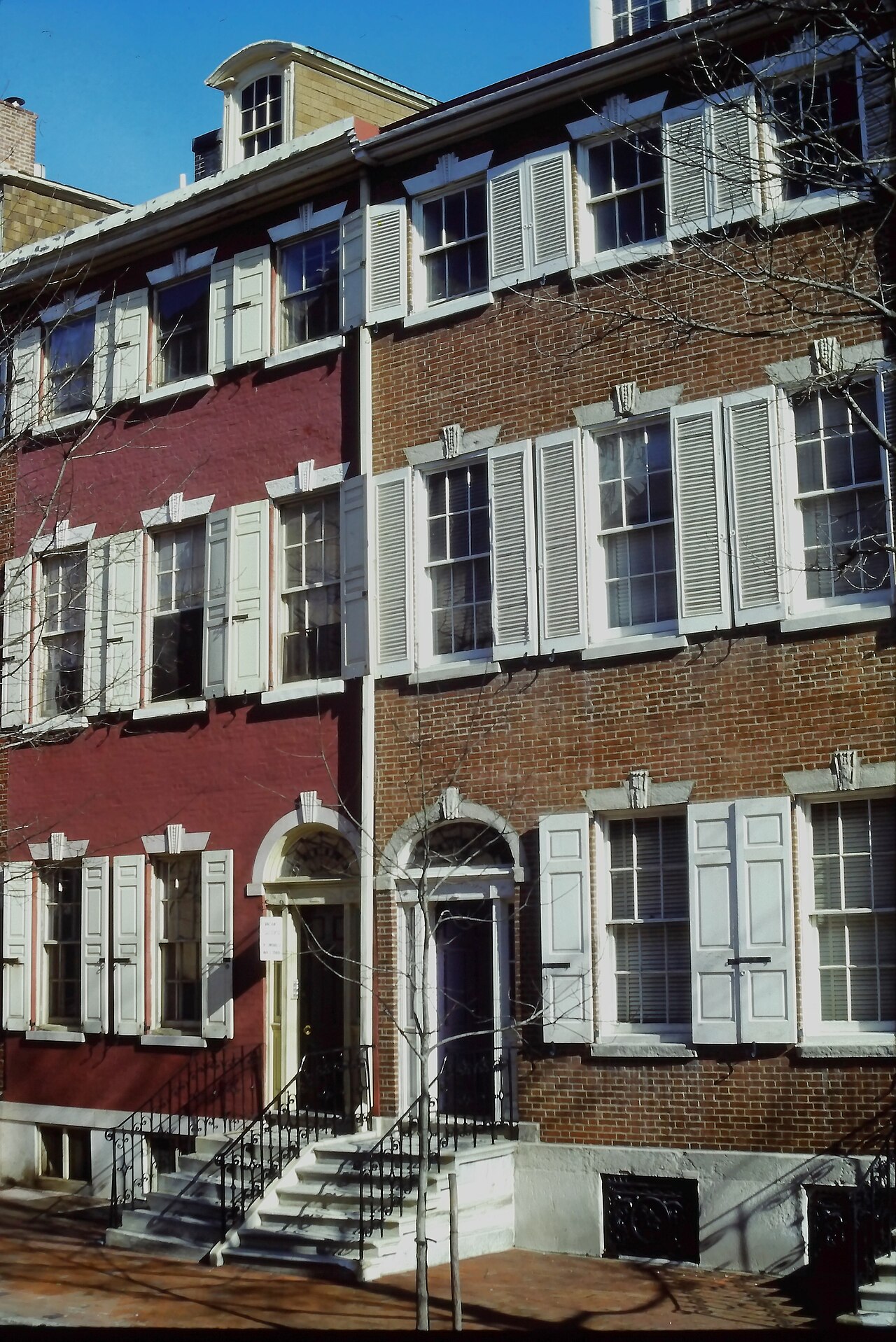 A pair of Federal rowhouses in red brick, one painted a deep red, arched doorways side by side under a shared cornice line, white shuttered windows on all three storeys.