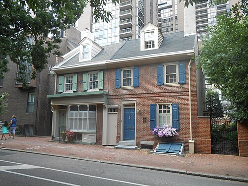 A small pair of colonial houses on Spruce Street, blue shutters and a blue front door, dormered windows and a brick sidewalk, modern glass towers rising directly behind.
