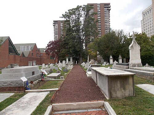 Old St. Mary's churchyard on Fourth Street, rows of white grave markers along a gravel path, a brick perimeter wall, tall apartment towers behind the trees.