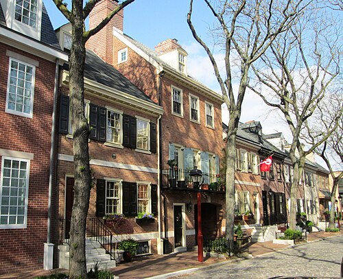 A pair of Delancey Street rowhouses, marble front steps, a wrought iron balcony over a garden gate, dormered gables above and a cobblestone lane at the curb.