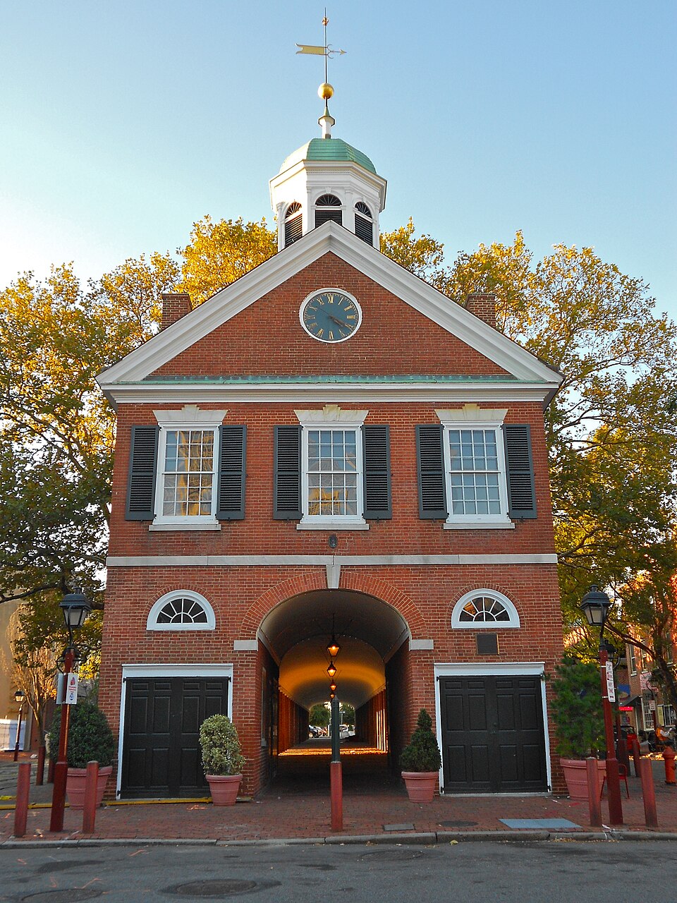 The Head House market shed at Second and Pine, a white octagonal cupola over a covered brick arcade, garage doors on either side, potted plants and red bollards along the curb.