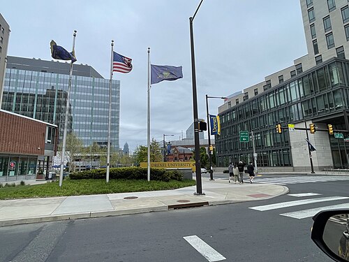 The Drexel University sign and flagpoles at a street corner, glass academic buildings behind and pedestrians crossing at the light.