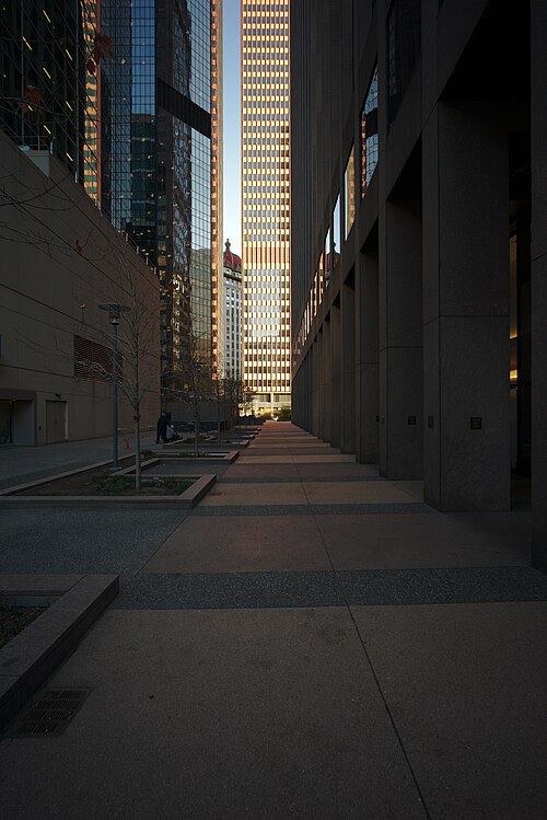 A narrow granite paved walkway between dark stone office towers, glass curtain walls reflecting a taller tower and a green domed roof beyond.