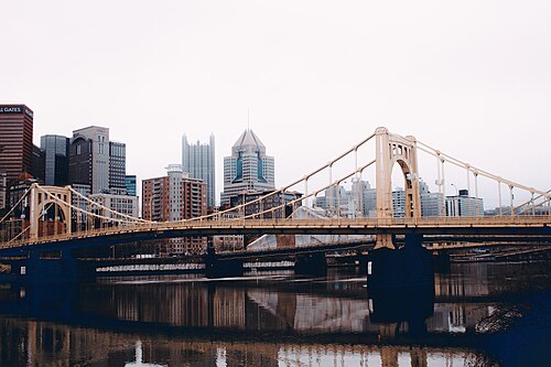 A second yellow self-anchored suspension bridge over the Allegheny in daylight, another identical span visible downstream and PPG Place's glass spires behind.