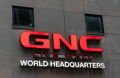 GNC's illuminated red and white headquarters signage mounted on a grey stone office tower facade, two dark windows above it.