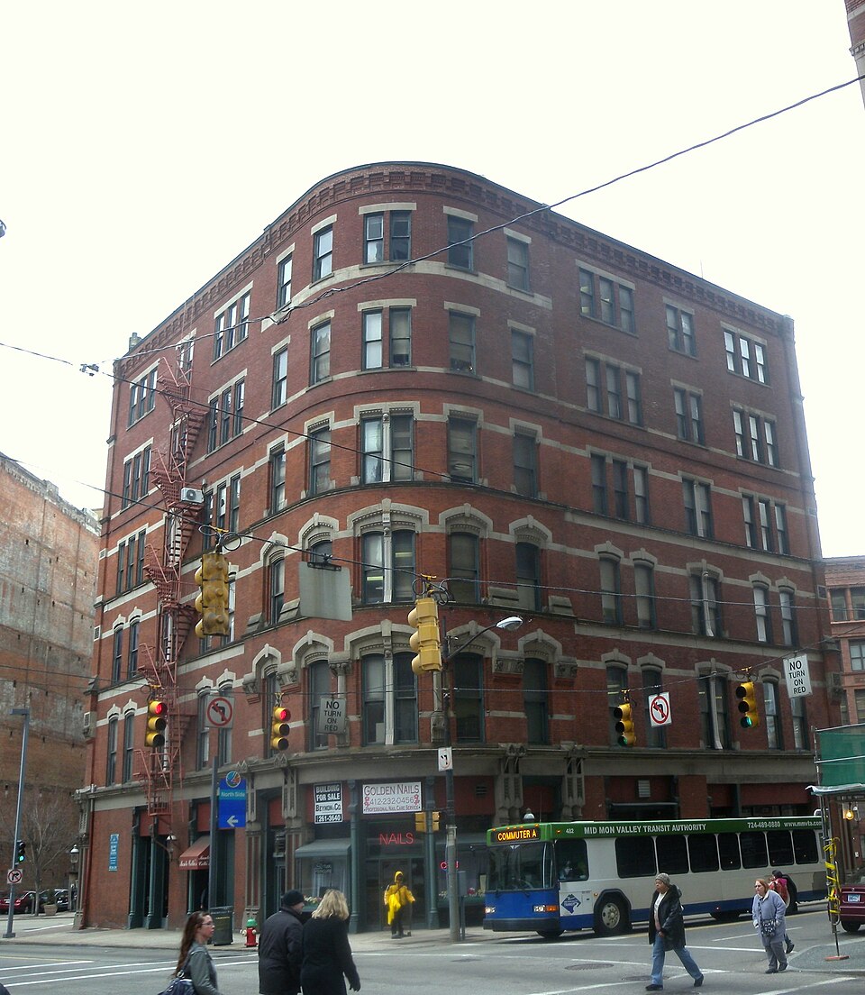 A curved six storey red brick commercial building at Seventh Avenue and Smithfield Street, traffic signals and a transit bus passing.