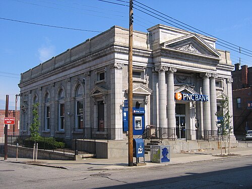 A stone bank building with illuminated PNC Bank channel letters at a paved Butler Street corner, an ATM and iron fencing along the front.
