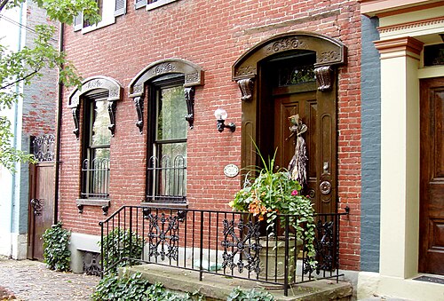 A red brick rowhouse facade with carved stone window and door surrounds, wrought iron railings, potted plants and ivy at the front steps.