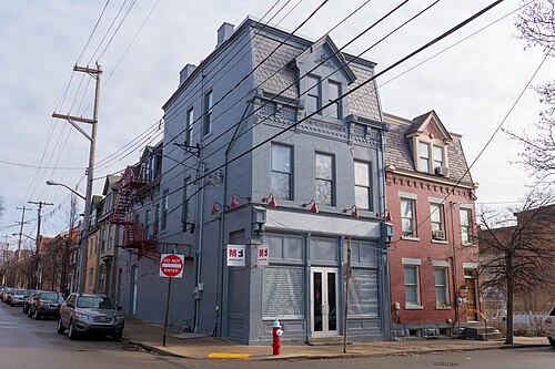 A grey painted three storey corner building with a mansard roof and fire escape at Monterey Street, a red brick Victorian rowhouse beside it and more rowhouses receding down the block.