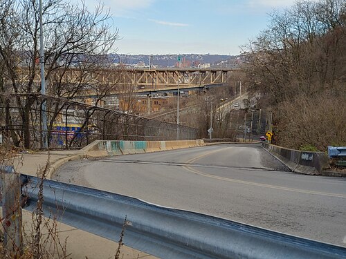 A two lane street curving downhill past metal guardrails and chain link fencing, bridges visible in the valley below.