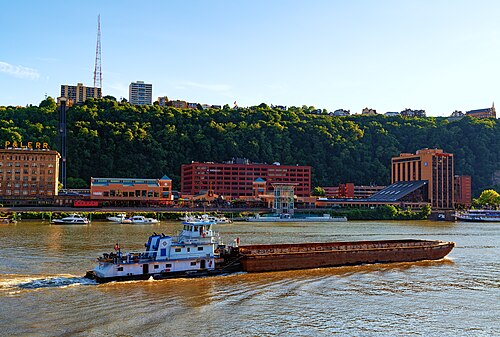A tugboat pushing a barge on the river below the wooded cliff, houses along the top of the ridge.