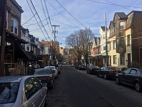 Atwood Street narrowing between brick storefronts and rowhouses, with cars parked under utility wires.