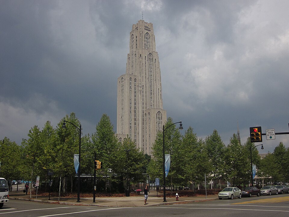 The Cathedral of Learning tower rising above green trees around a lawn and street intersection, confirmed against the scene's own Street View frame.