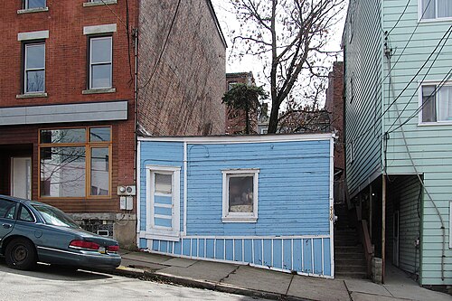 A small pale blue wood-frame house squeezed between a red brick building and a mint green sided building, with city steps visible alongside it.