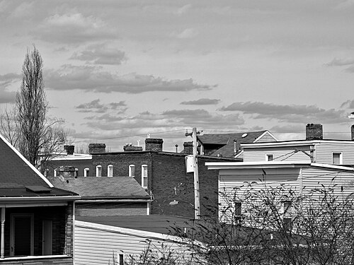 A black and white view of tightly packed rooflines, chimney pots and brick and wood-frame gables above a residential block.