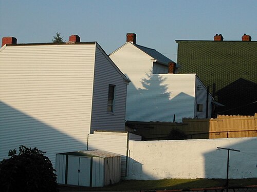 The blank side and rear walls of wood-frame houses in white and green siding above a low painted yard wall and a metal storage shed.