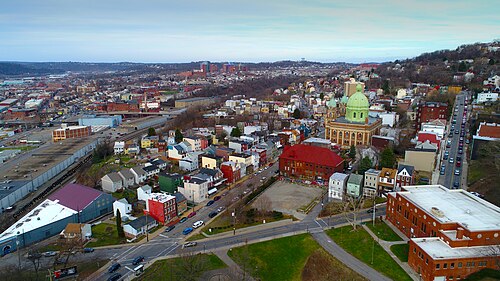 An aerial view over the hillside from West Penn Park, the green domed church at the center, rowhouse rooftops on the slope and a rail corridor in the valley below.