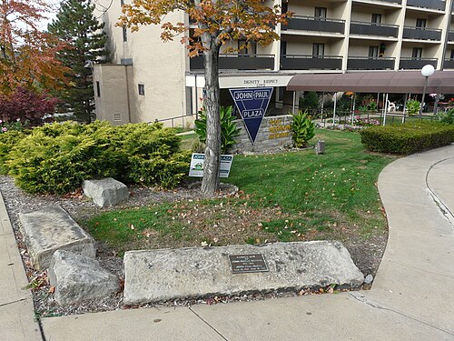 John Paul Plaza, a beige brick apartment building on Herron Avenue with a stone entrance sign and a landscaped lawn.