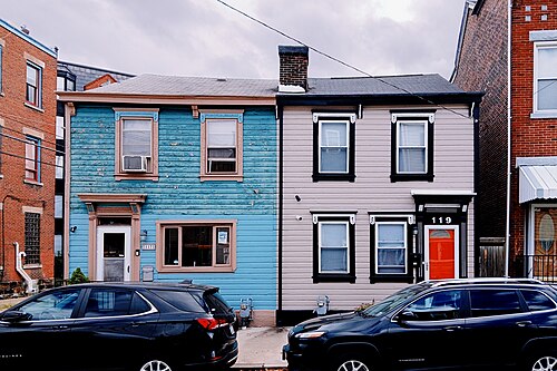 Two adjoining wood sided rowhouses on South 24th Street, one painted blue and one tan, black cars parked at the curb.