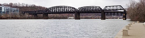 The Hot Metal Bridge, a black steel truss bridge crossing the Monongahela River, a paved riverside path in the foreground.