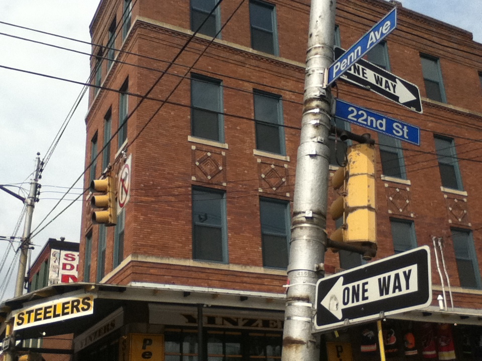 A four storey brick corner building at Penn Avenue and 22nd Street, diamond brickwork above the windows, a Yinzers storefront and awning below, street signs and traffic signals at the corner.
