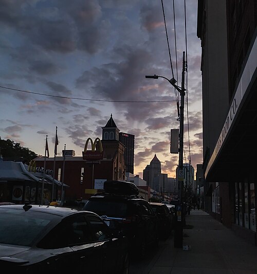 A street corner at dusk with a lit McDonald's sign since closed and demolished, a painted food company awning, parked cars along the curb and downtown Pittsburgh's skyline behind.