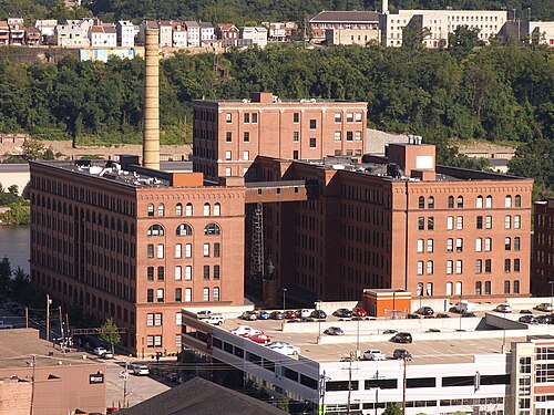 The Armstrong Cork Factory complex seen from across the river, a multi storey red brick warehouse with a tall chimney stack and a skybridge between wings.