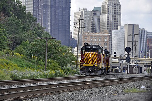 A freight locomotive on the riverside tracks with downtown Pittsburgh's towers rising behind it.