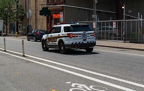 A police SUV on Penn Avenue beside a plain warehouse wall, a chain link fenced lot and a hanging shamrock sign.