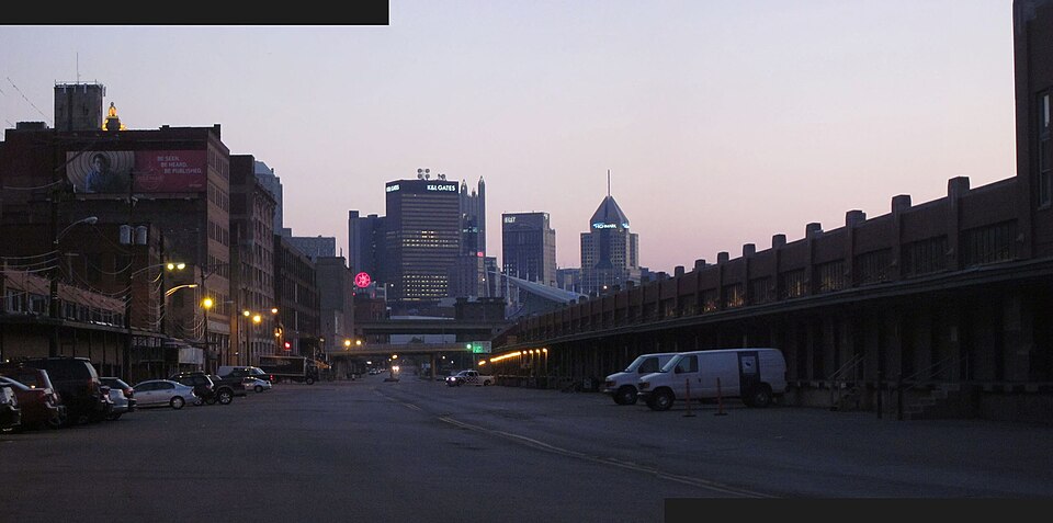 A produce warehouse's canopied loading dock running the length of a block on Smallman Street, delivery vans backed against it and the downtown skyline behind, photographed at dusk before the terminal's current renovation.