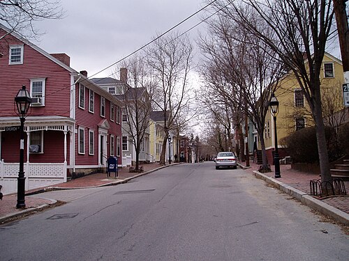 A colonial street of red, yellow and gray clapboard houses close along the sidewalk, with gas lamp posts, brick paving at the curbs and a single overhead wire.