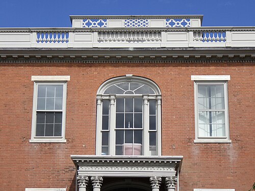 The upper facade of a red brick Federal house, a rounded window with fluted columns over the entrance and a roof balustrade with an open fretwork pattern.