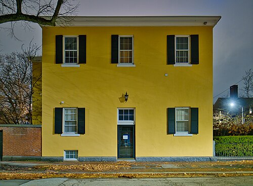 A yellow stucco Federal house at night, black shutters, a granite foundation and a single lantern lit over the front door.