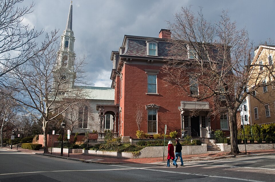 A red brick Victorian mansion with a mansard roof and bracketed cornice on Benefit Street, the First Unitarian Church's white steeple rising behind it, two people crossing the brick crosswalk.