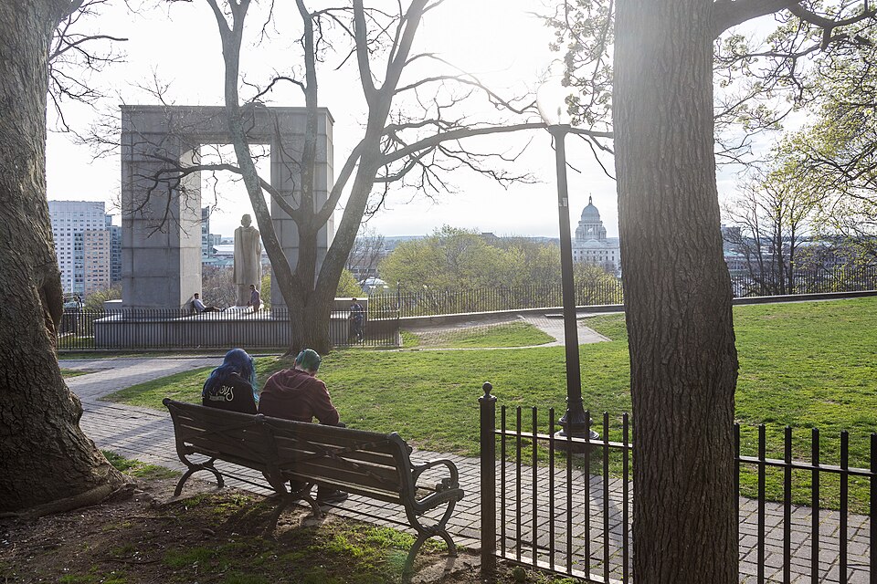 A granite monument arch framing the Roger Williams statue in Prospect Terrace Park, an iron fence and green lawn, with the State House dome and downtown Providence visible through the trees.
