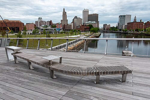 A wooden pedestrian bridge over the river with the downtown skyline behind it, the Industrial Trust tower and a domed building visible past the trees.