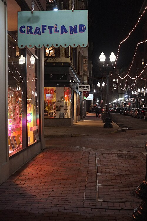 A lit shop sign over a brick sidewalk on a downtown street at night, string lights strung between buildings and storefronts glowing along the block.