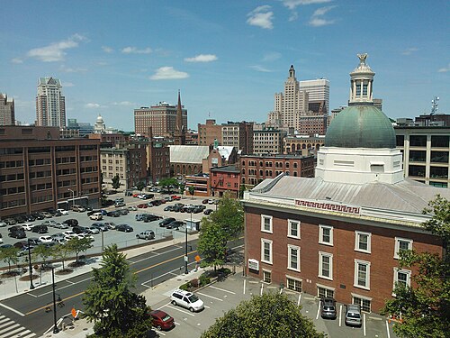A high angle view over a brick church with a domed cupola toward the downtown skyline, the State House dome and office towers visible past a parking lot.