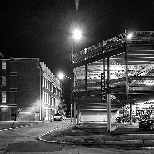 A multi level steel parking structure on a downtown corner at night, a lit PARK sign over the entrance and a brick building alongside.